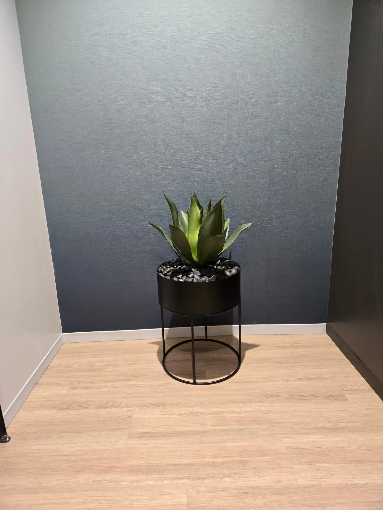 Faux agave in a round black planter on a metal stand, set against a navy textured wall in an alcove